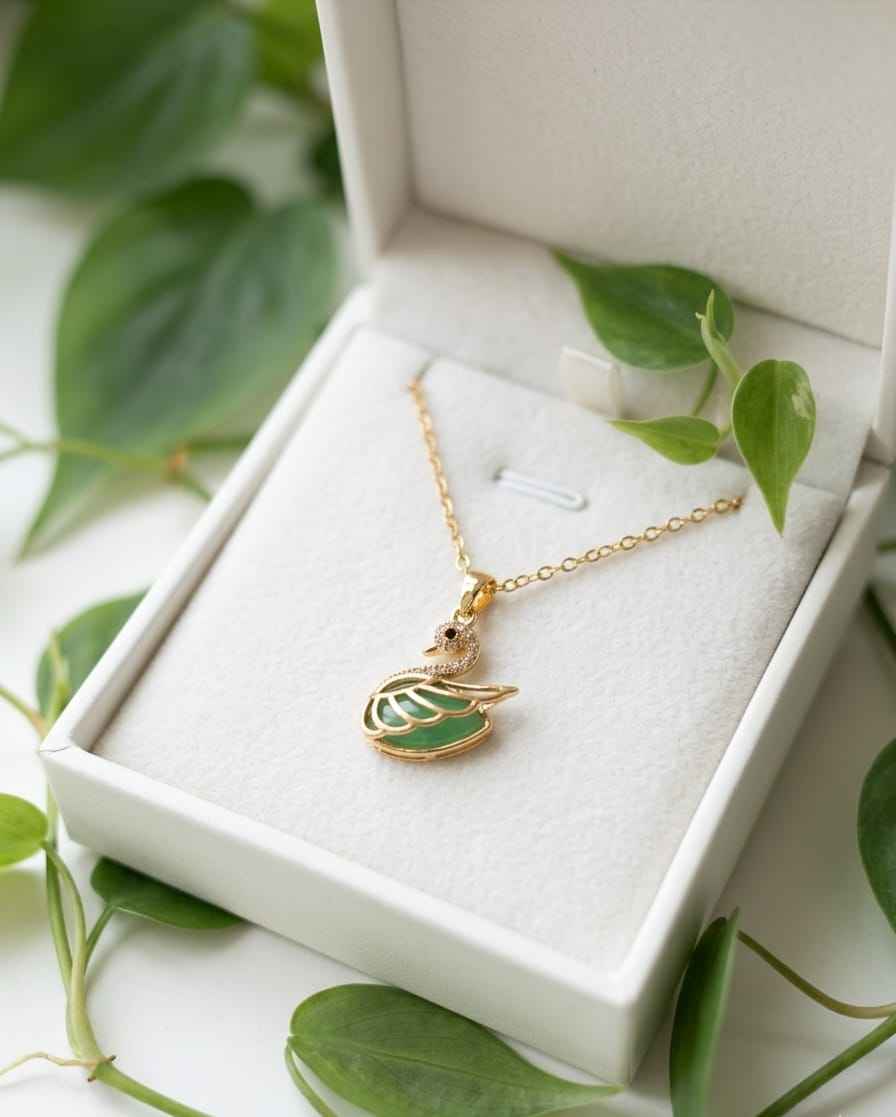 Gold swan pendant necklace with green enamel, shown in a gift box on white fabric.